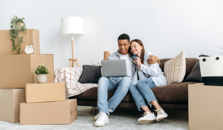 Real Estate Concept. Happy Mixed Race Family Couple Sitting On A Sofa Between Cardboard Boxes In Living Room Of Their New Home, Using Laptop And Credit Card For Online Shopping, Buying Things For Home