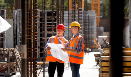 Construction Manager And Engineer Dressed In Orange Work Vests And Hard Helmets Explore Construction Documentation On The Building Site Near The Steel Frames