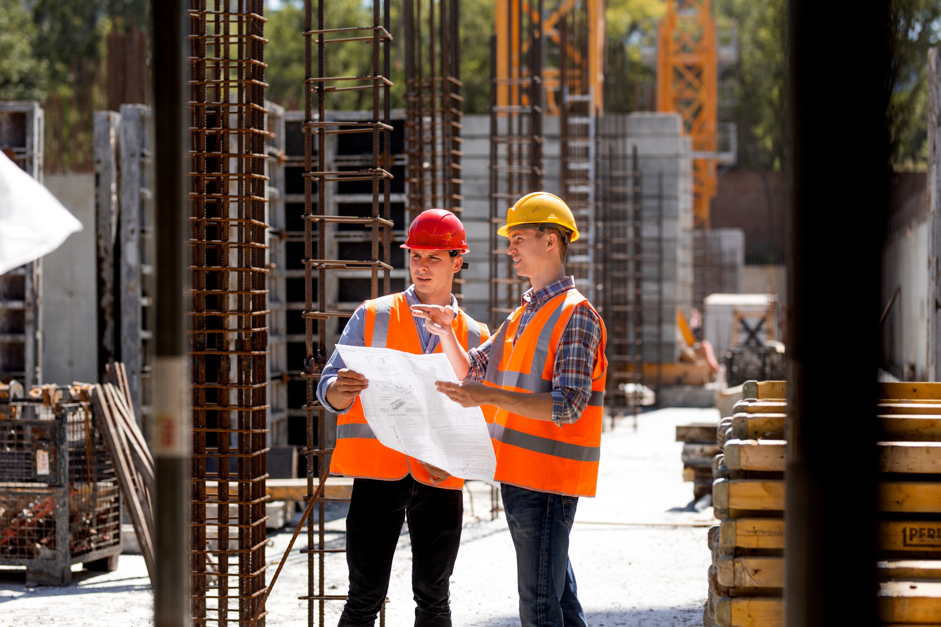 Construction Manager And Engineer Dressed In Orange Work Vests And Hard Helmets Explore Construction Documentation On The Building Site Near The Steel Frames