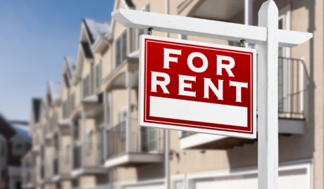 For Rent Real Estate Sign In Front Of A Row Of Apartment Condominiums Balconies And Garage Doors.